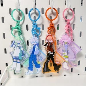 Peut inclure: Quatre porte-clés en acrylique représentant des personnages de style anime avec des palettes de couleurs distinctes. Chaque breloque est suspendue à un anneau et une chaîne en plastique assortis. Les personnages portent des tenues variées.