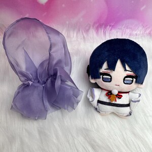 May include: A plush doll with a dark blue hair and a white outfit with black accents. A purple sheer hood is next to the doll. The doll has a red bow and a small leaf-shaped charm.