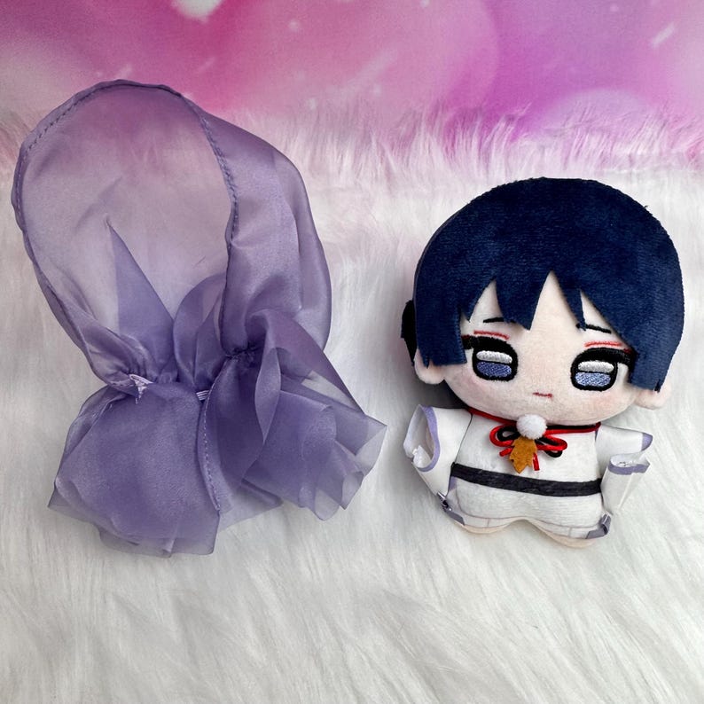May include: A plush doll with a dark blue hair and a white outfit with black accents. A purple sheer hood is next to the doll. The doll has a red bow and a small leaf-shaped charm.