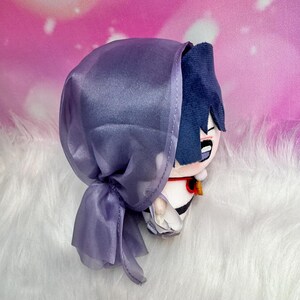 May include: A plush doll with blue hair and a purple hood. The doll has a white and black outfit with a red and gold accent. The doll is on a white, fluffy surface.