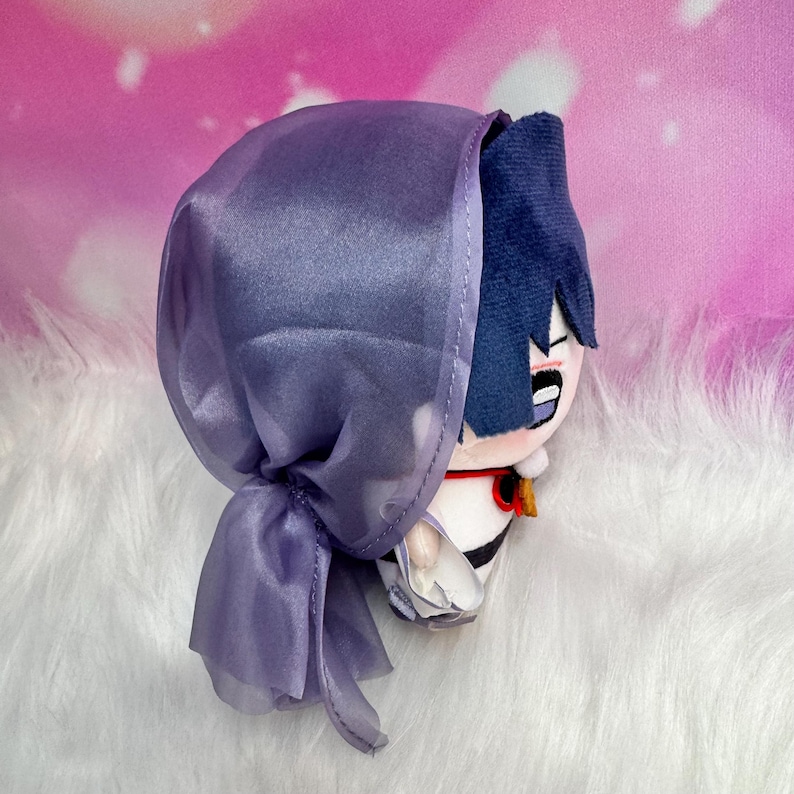 May include: A plush doll with blue hair and a purple hood. The doll has a white and black outfit with a red and gold accent. The doll is on a white, fluffy surface.
