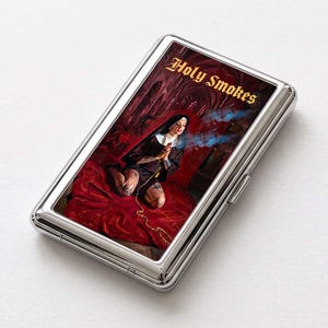 May include: A rectangular silver metal cigarette case featuring a graphic print of a person dressed in a nun's habit kneeling on a red surface. The image includes blue smoke and the text Holy Smokes in a gothic font style at the top.