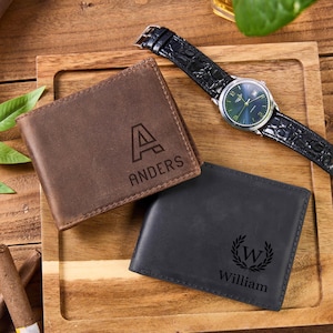 May include: A brown leather wallet with the name "Anders" and initial "A" engraved on it, and a black leather wallet with the name "William" and a laurel wreath engraved on it. A wristwatch with a black leather band is also present.