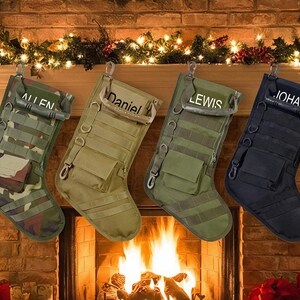 Könnte beinhalten: Vier taktische Weihnachtsstrümpfe hängen über einem Kamin. Die Strümpfe sind in Grün-, Braun- und Schwarztönen gehalten und tragen Namen wie "ALLEN", "Daniel", "LEWIS" und "JOHAN". Sie haben mehrere Taschen und Riemen.