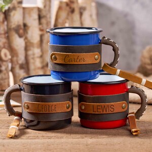 Puede incluir: Tres tazas de esmalte en azul, negro y rojo, cada una con una envoltura y asa de cuero. La taza azul tiene "Carter" en el cuero, la taza negra "George" y la taza roja "Lewis". Las tazas est&aacute;n sobre una superficie de madera.