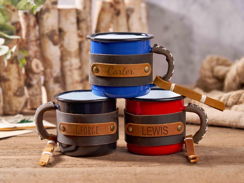 Puede incluir: Tres tazas de esmalte en azul, negro y rojo, cada una con una envoltura y asa de cuero. La taza azul tiene "Carter" en el cuero, la taza negra "George" y la taza roja "Lewis". Las tazas est&aacute;n sobre una superficie de madera.
