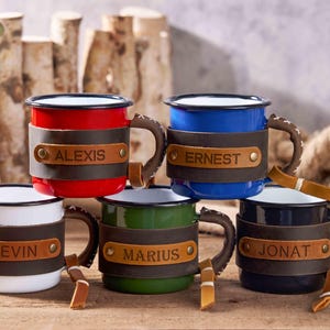 Puede incluir: Un conjunto de tazas de esmalte en varios colores, como rojo, azul, blanco, verde y negro. Cada taza tiene una envoltura de cuero con un nombre, como "Alexis" o "Ernest". Las tazas tienen asas marrones y se exhiben con detalles de madera.