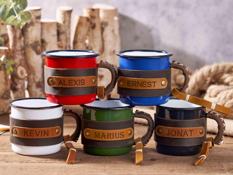 Puede incluir: Un conjunto de tazas de esmalte en varios colores, como rojo, azul, blanco, verde y negro. Cada taza tiene una envoltura de cuero con un nombre, como "Alexis" o "Ernest". Las tazas tienen asas marrones y se exhiben con detalles de madera.