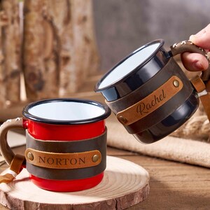 Puede incluir: Dos tazas de esmalte personalizadas. Una es roja con "NORTON" en una banda de cuero, la otra es negra con "Rachel" en una banda de cuero. Ambas tienen asas y detalles de cuero marr&oacute;n. Las tazas est&aacute;n sobre una superficie de madera.