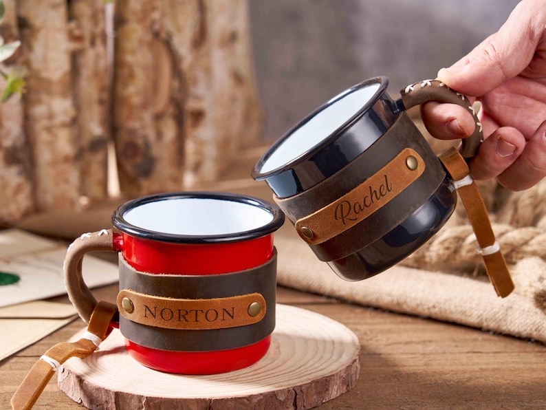 Puede incluir: Dos tazas de esmalte personalizadas. Una es roja con "NORTON" en una banda de cuero, la otra es negra con "Rachel" en una banda de cuero. Ambas tienen asas y detalles de cuero marr&oacute;n. Las tazas est&aacute;n sobre una superficie de madera.