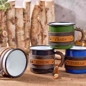 Puede incluir: Tazas de camping de esmalte en verde, azul, negro y blanco con detalles de cuero. Cada taza tiene una banda de cuero con un nombre: "Chester", "Edison", "Tobias" y "Samuel". Las tazas tienen asas cosidas marrones y est&aacute;n sobre madera.