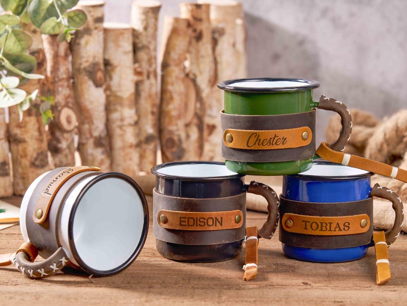 Puede incluir: Tazas de camping de esmalte en verde, azul, negro y blanco con detalles de cuero. Cada taza tiene una banda de cuero con un nombre: "Chester", "Edison", "Tobias" y "Samuel". Las tazas tienen asas cosidas marrones y est&aacute;n sobre madera.