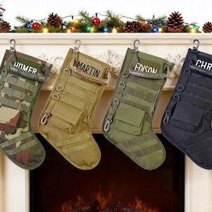May include: Four tactical Christmas stockings in camouflage, olive green, and black hang from a fireplace mantel. Each stocking has a name printed on it: Homer, Martin, Edison, and Chris. The mantel is decorated with Christmas lights and pinecones.