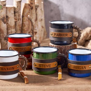 Puede incluir: Una colecci&oacute;n de tazas de esmalte personalizadas en varios colores: blanco, rojo, verde, azul y negro. Cada taza presenta una envoltura de cuero con un nombre y un asa cosida. Las tazas se exhiben sobre una superficie de madera con un fondo r&uacute;stico.
