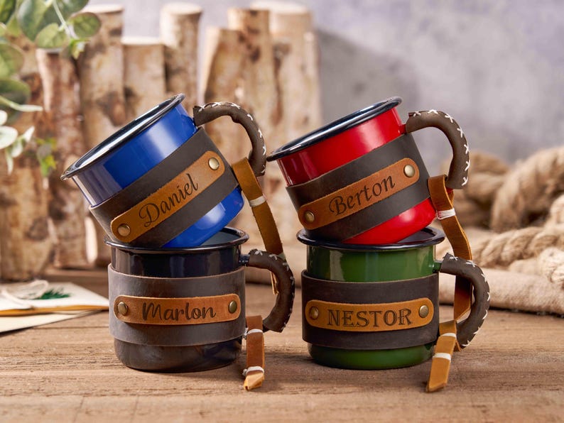 Puede incluir: Cuatro tazas de esmalte personalizadas en azul, rojo, negro y verde. Cada taza tiene una envoltura de cuero con un nombre grabado. Las asas est&aacute;n envueltas en cuero marr&oacute;n con costuras blancas. Las tazas est&aacute;n apiladas sobre una superficie de madera.