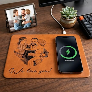 Op de afbeelding: Een bruin lederen draadloos oplaadpad met een familieportret en de tekst "We love you!". Een smartphone wordt op het pad opgeladen en toont een groen oplaadsymbool. Een ingelijste familiefoto is ook aanwezig.