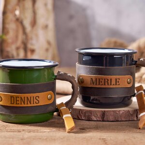 Puede incluir: Dos tazas de esmalte personalizadas, una verde y otra negra. Cada taza tiene una envoltura de cuero con un nombre. Las asas son marrones con un dise&ntilde;o cosido. Las tazas est&aacute;n sobre una superficie de madera.