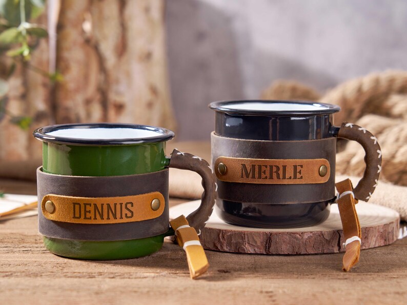 Puede incluir: Dos tazas de esmalte personalizadas, una verde y otra negra. Cada taza tiene una envoltura de cuero con un nombre. Las asas son marrones con un dise&ntilde;o cosido. Las tazas est&aacute;n sobre una superficie de madera.