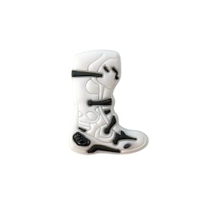 Puede incluir: Un dije de bota de motocross blanca y negra. La bota presenta correas y detalles en negro, con un diseño detallado. El dije está diseñado para parecerse a una bota protectora utilizada en los deportes de motor. La bota es un solo artículo, sobre un fondo blanco.