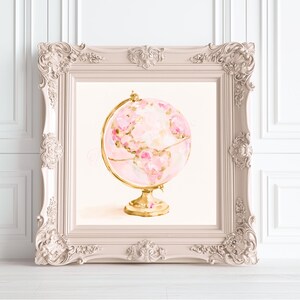 Puede incluir: Un globo terráqueo decorativo con una esfera de color rosa pálido con patrones florales abstractos en tonos rosa y blanco, sostenido por un soporte y arco meridiano de metal dorado, dentro de un marco rectangular claro y ornamentado.