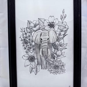 May include: Black framed art print featuring a detailed pen and ink drawing of an elephant surrounded by flowers. The elephant is shaded in gray, with the flowers and leaves in black and white. The artwork is displayed in a black wooden frame.
