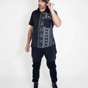 May include: A black short-sleeved shirt with a white geometric pattern. The shirt has a button-up collar and a pocket on the left side. The person is wearing black pants and black boots.