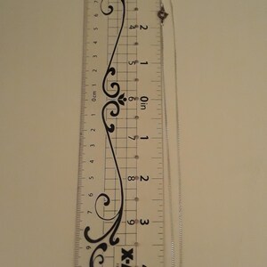 May include: A silver necklace with a Mickey Mouse pendant, next to a ruler. The ruler is marked in inches and centimeters. The necklace is approximately 6 inches long. The pendant has clear stones.