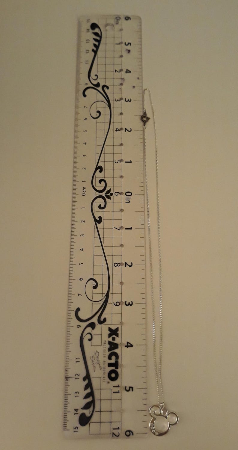 May include: A silver necklace with a Mickey Mouse pendant, next to a ruler. The ruler is marked in inches and centimeters. The necklace is approximately 6 inches long. The pendant has clear stones.