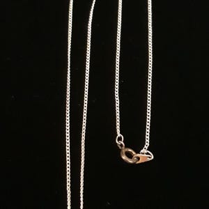 May include: Silver-toned necklace with a Mickey Mouse-shaped pendant. The pendant has a curved design with three clear, round stones. The chain is a delicate, linked style. The background is solid black.
