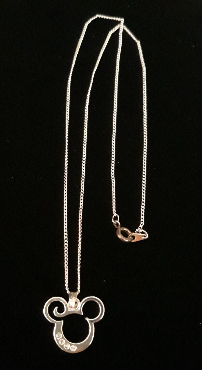 May include: Silver-toned necklace with a Mickey Mouse-shaped pendant. The pendant has a curved design with three clear, round stones. The chain is a delicate, linked style. The background is solid black.
