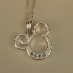 May include: A silver-colored pendant necklace featuring a Mickey Mouse silhouette. The pendant has a smooth, polished finish and is adorned with four small, clear gemstones. The necklace chain is also silver and delicate.