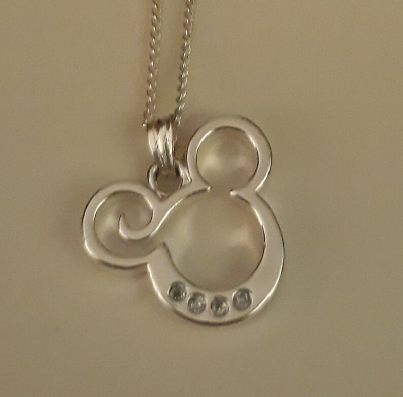 May include: A silver-colored pendant necklace featuring a Mickey Mouse silhouette. The pendant has a smooth, polished finish and is adorned with four small, clear gemstones. The necklace chain is also silver and delicate.
