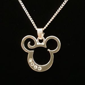 May include: A silver-colored Mickey Mouse pendant necklace. The pendant features the iconic Mickey Mouse head shape with three small, clear gemstones embedded in the lower portion. The necklace chain is a delicate, linked design, and the background is a solid black.