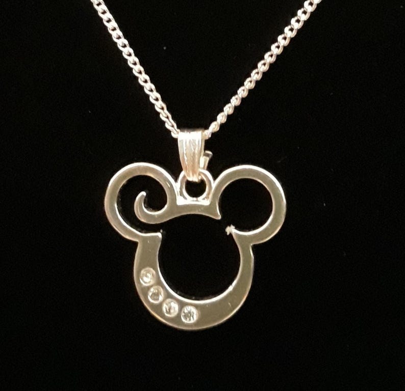 May include: A silver-colored Mickey Mouse pendant necklace. The pendant features the iconic Mickey Mouse head shape with three small, clear gemstones embedded in the lower portion. The necklace chain is a delicate, linked design, and the background is a solid black.