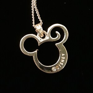 May include: A silver-colored pendant necklace featuring the iconic Mickey Mouse silhouette. The pendant is a cutout design with the word "DISNEY" inscribed on the bottom. The necklace hangs on a delicate silver chain against a black background.