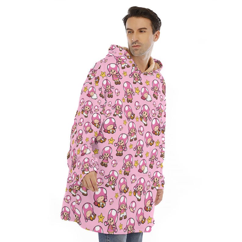 Puede incluir: Manta con capucha rosa con un patr&oacute;n repetido de un personaje de dibujos animados con cabeza de hongo. La manta tiene un interior suave y afelpado y una capucha. El personaje lleva un vestido rosa y sostiene una estrella. La manta est&aacute; dise&ntilde;ada para la comodidad y el calor.