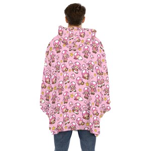 Puede incluir: Sudadera con capucha rosa con un patr&oacute;n repetido de un personaje de dibujos animados con una gorra de hongo rosa, estrellas y otros elementos. La sudadera es usada por una persona con jeans azules.