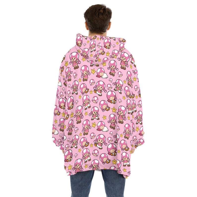 Puede incluir: Sudadera con capucha rosa con un patr&oacute;n repetido de un personaje de dibujos animados con una gorra de hongo rosa, estrellas y otros elementos. La sudadera es usada por una persona con jeans azules.