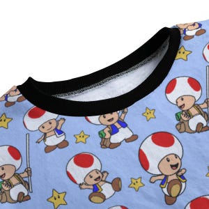 May include: Light blue sweatshirt featuring a repeating pattern of the character Toad from the Super Mario video game series. Toad has a white mushroom cap with red spots, a blue vest, and a tan backpack. Yellow stars are also scattered across the fabric.