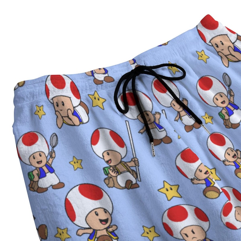 May include: Light blue shorts featuring a repeating pattern of a cartoon character with a red and white mushroom cap, blue vest, and brown shoes. The shorts have a black drawstring and yellow star accents. The fabric appears soft and comfortable.