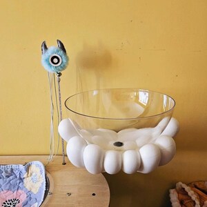 Puede incluir: Un cuenco blanco en forma de nube con un inserto de vidrio transparente cuelga del techo. Un juguete de peluche azul y blanco con cuernos está sujeto a una cuerda que cuelga del techo.