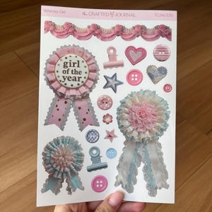 May include: A sheet of stickers with a whimsical theme. The stickers include rosettes, hearts, stars, buttons, and a paper clip. The text "Whimsy Girl" and "girl of the year" are visible.