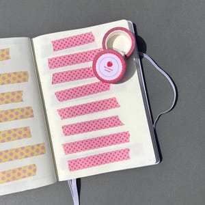May include: An open notebook with pink star-patterned washi tape strips and a roll of matching tape. The tape roll has a red and white label with a strawberry illustration and the text "The Crafted Journal". Yellow polka dot tape is also visible.