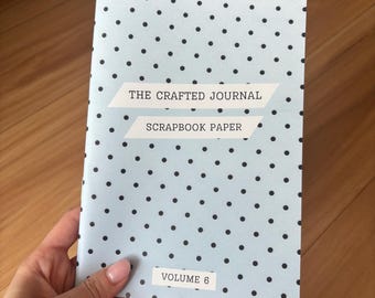 Może przedstawiać: Jasnoniebieski notatnik w czarne grochy. Okładka zawiera tekst "THE CRAFTED JOURNAL SCRAPBOOK PAPER" i "VOLUME 6". Notatnik jest trzymany w dłoni.