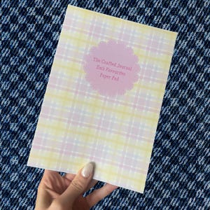May include: A rectangular paper pad featuring a soft yellow, white, and light pink plaid pattern. Centered on the cover is a scalloped pink circular label with the text The Crafted Journal Em's Favourites Paper Pad printed in a dark pink font.