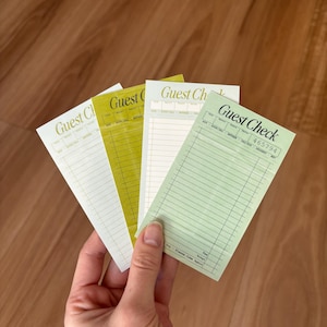 May include: A hand holds a fan of four guest check pads in shades of green and white. Each pad has the words "Guest Check" printed at the top. The pads have lines for writing and a space for the total.