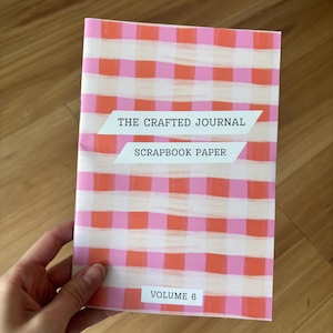 May include: A pink and orange gingham patterned journal titled "THE CRAFTED JOURNAL SCRAPBOOK PAPER" with "VOLUME 6" printed at the bottom. The journal is held in a person's hand.