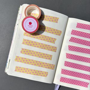 May include: An open journal with pages marked by decorative washi tape. Two rolls of tape are visible, one with a label that reads "The Crafted Journal" and features a red apple illustration. The tape on the pages has yellow and pink patterns.