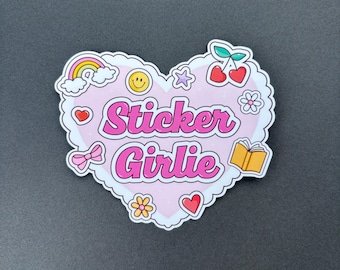 May include: A heart-shaped sticker with the words "Sticker Girlie" in pink script. The sticker is decorated with a rainbow, cherries, a smiley face, a star, a flower, a bow, a heart, and an open book. The sticker has a white border.