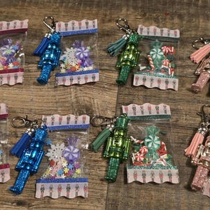 May include: A collection of Christmas-themed keychains. Each keychain features a miniature nutcracker figure, a tassel, and a clear bag filled with colorful candies and confetti. The keychains are in various colors, including pink, blue, and green.